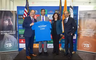 Three men and a woman stand shoulder-to-shoulder in front of a blue SUNY backdrop smiling and holding a blue t-shirt that reads "SUNY AI Symposium"