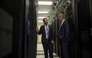 Two men in dark suits stand facing one another between two long rows of computer racks.