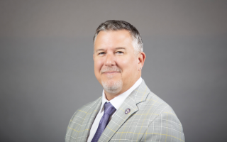 A man with short gray hair and a goatee wears a gray pinstripe suit and blue tie and smiles for a portrait against a gray backdrop.