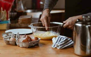 Various materials for cooking are arranged on a wooden kitchen countertop. A half-dozen egg carton is open and full of cracked eggshells. A man whisks the eggs in a glass bowl with bare hands.. 