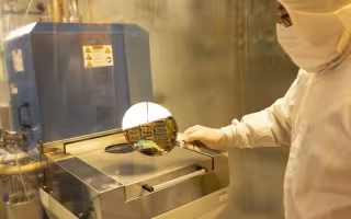 A man in a head-to-toe white cleanroom suit holds a silicon wafer while standing in front of a large metal-clad laboratory tool.