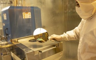 A man in a head-to-toe white cleanroom suit holds a silicon wafer while standing in front of a large metal-clad laboratory tool.