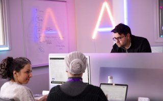 Two student sit facing in each other in front of a computer screen while a professor looks on with the letters "AI" illuminated on a wall behind them.