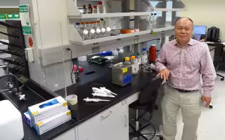 A man wearing a plaid button-down shirt and khaki pants poses, smiling, next to a lab bench covered in various pieces of chemistry equipment including pipettes, latex gloves, and plastic bottles containing clear liquids.
