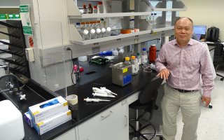 A man wearing a plaid button-down shirt and khaki pants poses, smiling, next to a lab bench covered in various pieces of chemistry equipment including pipettes, latex gloves, and plastic bottles containing clear liquids.