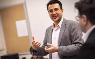 A man with short black hair and glasses in a gray blazer is pictured in a classroom.