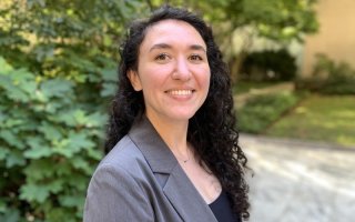 A young woman with dark curly hair wearing a gray blazer smiles for a portrait. Bushes and greenery are blurred in the background.