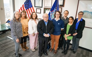 Eight people, including five women and three men smile together for a group portrait in an office with gray carpet and a collection of framed plaques in the background. An American flag and the flag of New York State stand behind the group.