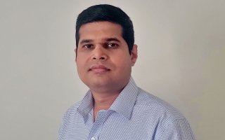 Headshot of a man with dark hair wearing a button down shirt with a blue and gray grid print, against a plain gray background.