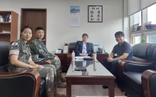 Two military personnel wearing uniforms are seated next to two civilians in an office. They smile and pose for a photo.