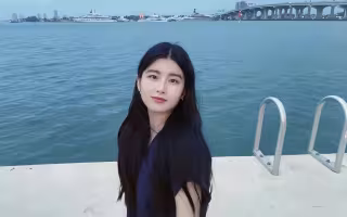 A young woman with long dark hair poses beside a marina. Blue water, a bridge and several large yachts are visible in the background. 