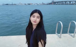 A young woman with long dark hair poses beside a marina. Blue water, a bridge and several large yachts are visible in the background. 