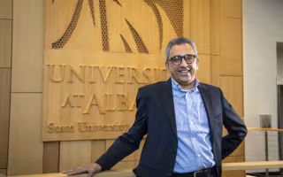 A man in a dark suit coat and button-down shirt smiles in front of a wood-paneled wall with the words "University at Albany."