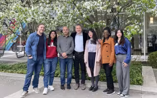 Seven people come together for a group photo outdoors. The group is standing in front of trees with white blossoms and the glass wall of UAlbany's lecture center concourse is behind them.
