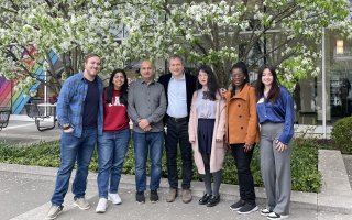 Seven people come together for a group photo outdoors. The group is standing in front of trees with white blossoms and the glass wall of UAlbany's lecture center concourse is behind them.