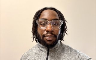 A young man with dark hair, short dark beard and glasses with square clear plastic frames smiles against a plain beige background.