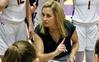 A woman with light brown hair and dressed in a black shirt is squated and pointing as several women's basketball players surround her in a team huddle on a basketball court.