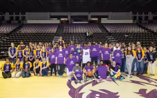 A large group of people wearing shirts with UAlbany written in all capital letters and a graphic of a Great Dane dog pose together on a basketball court with rows of empty seats behind them.