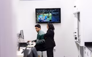 A man sits at desk looking at a computer screen while a woman stands behind him with her arms crossed.