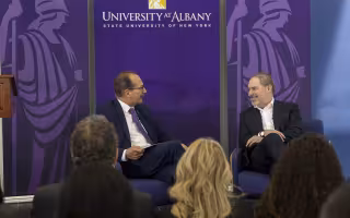 Two men are seated before a crowd of people in front of a purple backdrop with "University at Albany State University of New York" written on it with two Minerva logos adorning the sides.