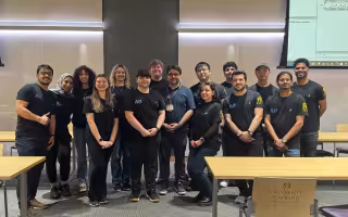 More than a dozen student researchers stand with Assistant Professor Abdullah Canbaz inside the ETEC shared training room.