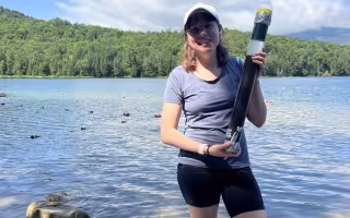 Sky Hooler holds up a sediment core sample in front of a body of water in the Adirondacks.
