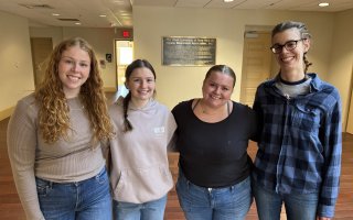 Four young women embrace, smiling, for a group photo in a hallway at UAlbany’s School of Social Welfare. They are dressed casually, all wearing blue jeans. The lighting is warm, with beige painted walls and wood floor. 