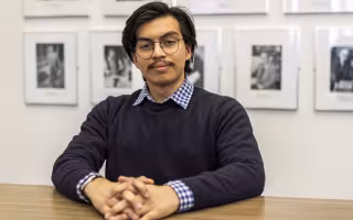 A young man with dark hair and round glasses, wearing a charcoal sweater, sits at a wooden desk with his hands folded, looking at the camera. The wall behind him is white and is hung with a series of black and white photos in equal-sized silver frames.