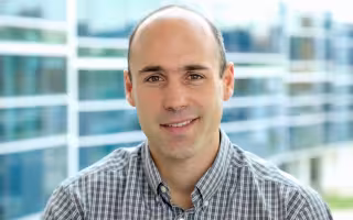 A man in a plaid shirt smiles for a portrait in front of a building with wavy glass