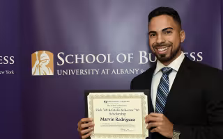A UAlbany student with dark hair and a beard stands with an award for a Scholarship with the School of Business sign behind him.