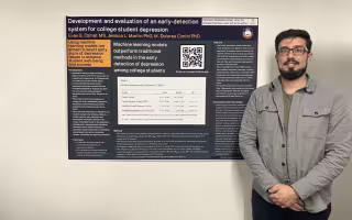 A young man with short brown hair and a goatee in black glasses, a gray button down shirt and dark pants stands against a wall next to a poster titled "Development and evaluation of an early-detection system for college student depression"