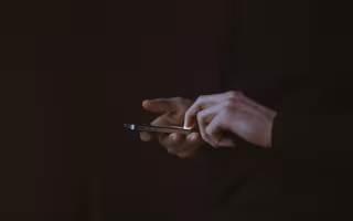 A pair of hands can be seen holding a phone in a dark room. The lit screen is the only source of light.