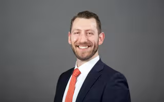 A man wearing a dark blue suit and dotted red tie with close-cropped brown hair and a full beard smiles at the camera in front of a gray background.