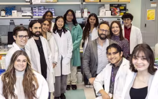 13 smiling people, including a mix of genders, in a science lab. Most are students wearing white lab coats. Shelves on the back wall hold boxes of lab supplies.