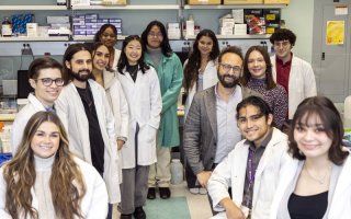 13 smiling people, including a mix of genders, in a science lab. Most are students wearing white lab coats. Shelves on the back wall hold boxes of lab supplies.