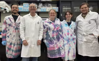 Five people, two women and three men, stand together smiling for a group portrait in front of a lab bench. All are wearing lab coats, three of which are tie dyed blue, pink and purple. Shelves of lab supplies can be seen behind them. 