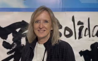 A woman with shoulder length blond hair smiles at the camera. She is wearing a pinstripe zip-up jacket and there is a banner behind her painted with Japanese characters.
