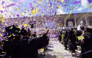 Purple and gold confetti rain down on a bunch of students standing in rows in front of chairs outdoors with the University at Albany podium in the distance against a blue sky with clouds.