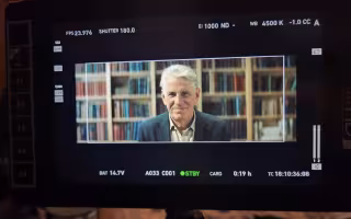A man with short white hair in a collared shirt and blazer appears on a film screen seated in front of wall-to-wall bookcases.