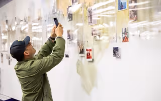 A man in a green jacket and dark cap takes a photo of a wall adorned with photos and artist renderings of UAlbany veterans