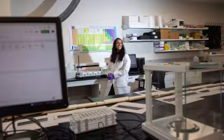 A smiling woman in a white lab coat and purple safety gloves stands in a laboratory.