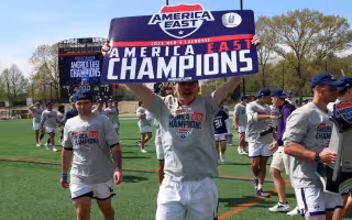 A man in a gray shirt and white shorts holds a sign that says America East Champions as his teammates walk behind him on a green athletic field during the day.