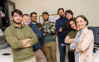 A group of eight people (two women and six men) stand together in a lab space with shelves on one side and a bank of computers on the other. All are smiling, facing the camera, with their arms folded across their chests. 