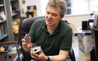 A a seated man in a green polo shirt leans toward the camera while holding a small, rectangular metallic box.