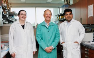 Three people, one woman and two men, stand together for a group portrait in a science lab. All are smiling and wearing lab coats.