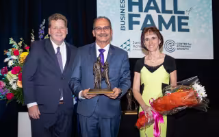 Three people stand smiling in front of a "Business Hall of Fame" sign, the man in the center holding a trophy depicting thee people, and the woman on the right holding flowers.