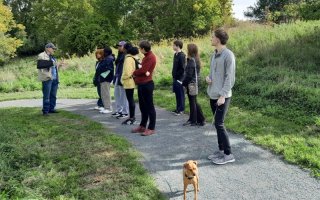 A group of students standing along a nature path listen as a man in a sling and ballcap talks. One students holds a dog on a leash.