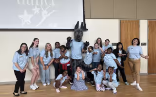 A group of children and adults in light blue shirts gather around Damien, UAlbany's Great Dane mascot, and pose for a celebratory picture in front of a screen that says "dance"
