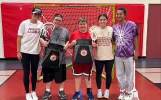 A group of three women wearing Lacrossing Barriers shirts stand with two men with special needs in front of a lacrosse net inside of a gym. A woman at the far right of the photo wears a UAlbany lacross jersey.