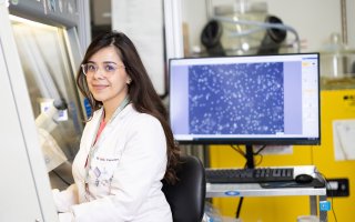 A young woman with dark hair and glasses wearing a white lab coat sits at a research station under a hood. Behind her is a monitor with a blue screen and tiny heart cells on display.
